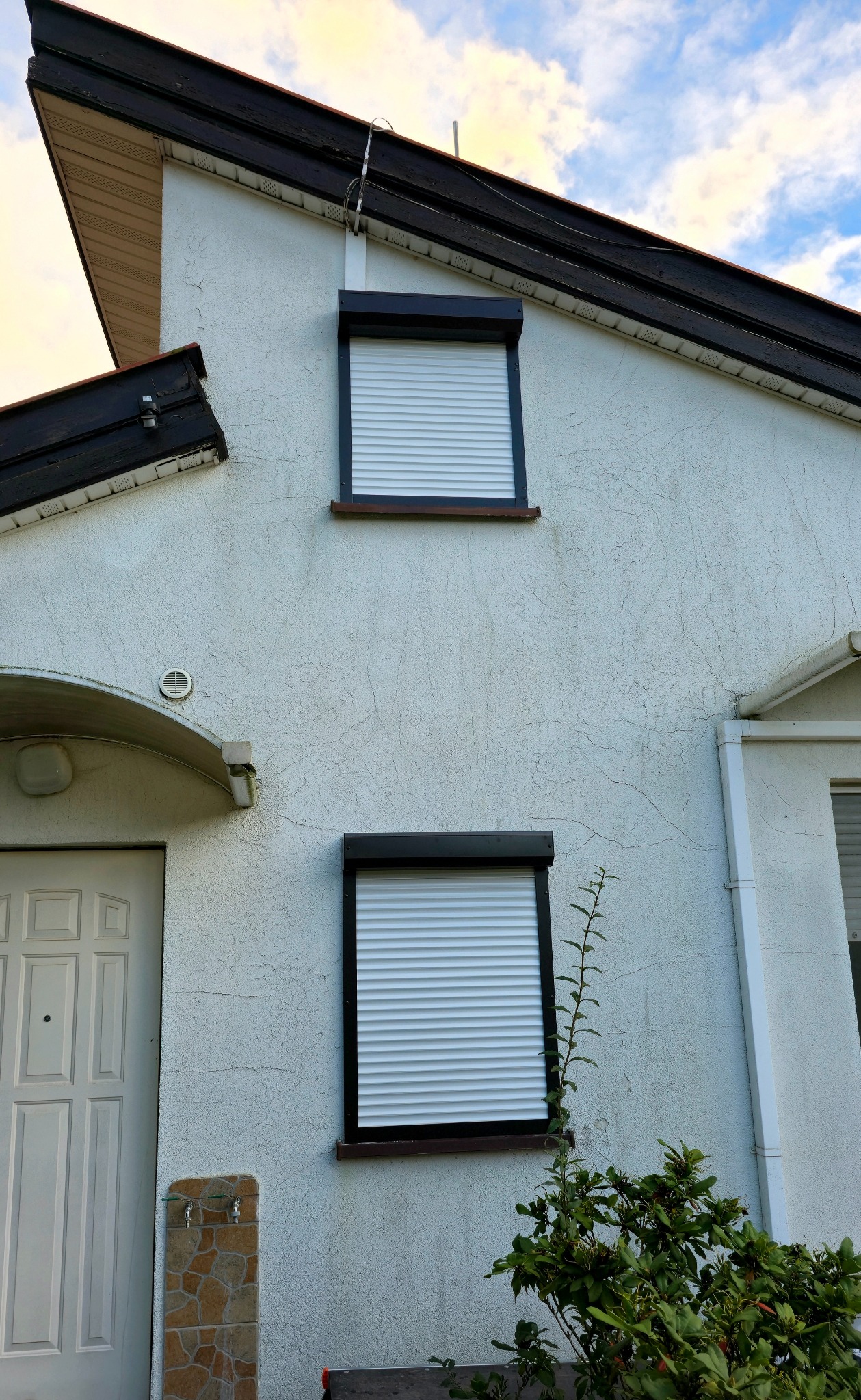 Biały dom z dwoma oknami z opuszczonymi, białymi  roletami zewnętrznymi w czarnych ramach. Obok dolnego okna rośnie zielony krzew.