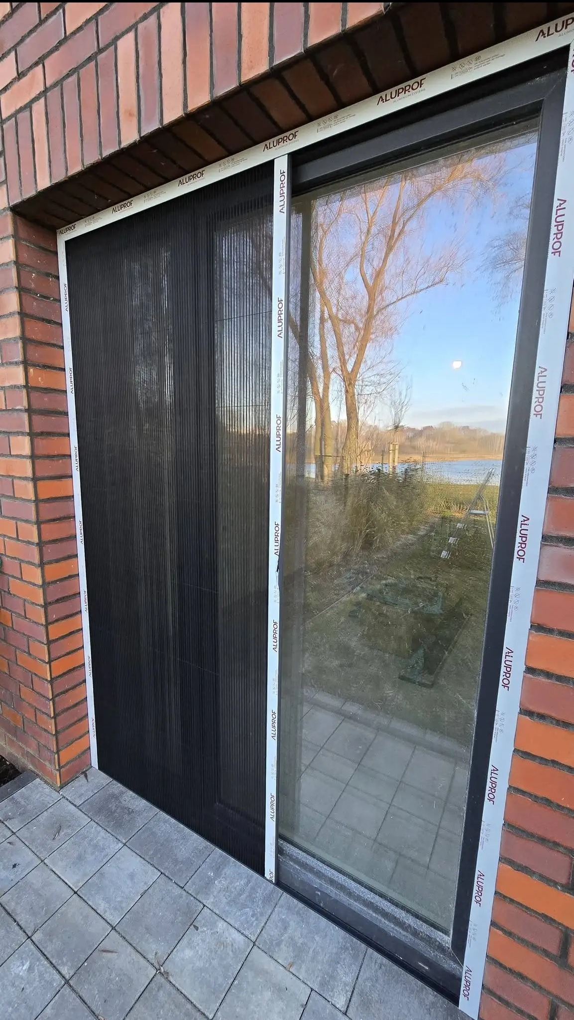 Ciemna plisa okienna w aluminiowej ramie, zamontowana w oknie budynku z cegły. Widok na zewnątrz przez szybę, częściowo zasłonięty.