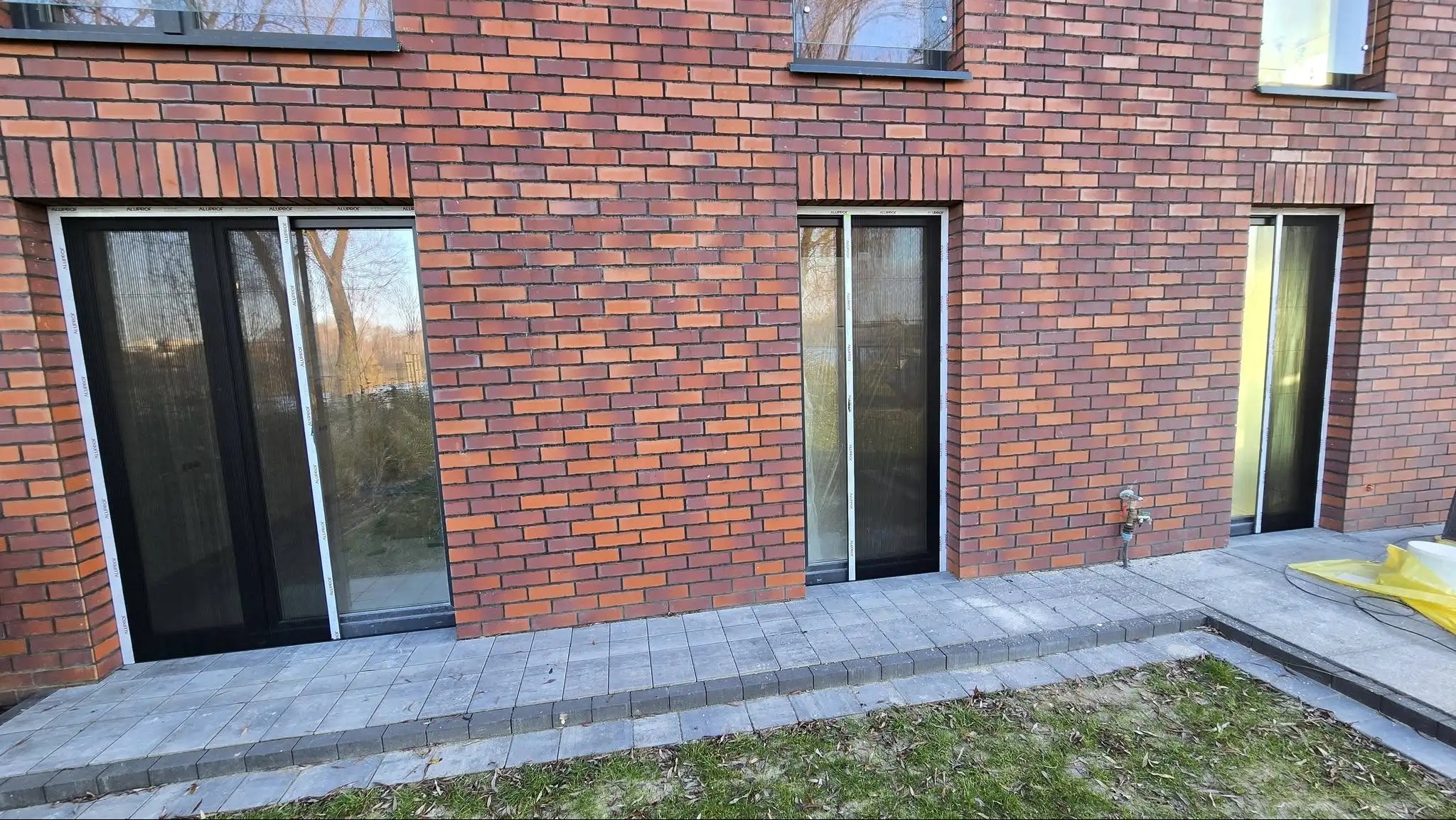 Ceglana elewacja budynku z trzema oknami zabezpieczonymi moskitierami w białych ramach. Widoczne detale wykończenia i fragment chodnika.