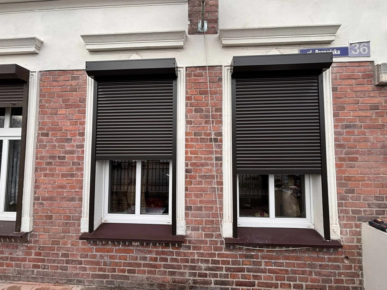 Ciemne rolety zewnętrzne na oknach w ceglanym budynku, widoczny fragment ulicy z numerem 36, białe ramy okienne i parapety w kolorze brązowym.