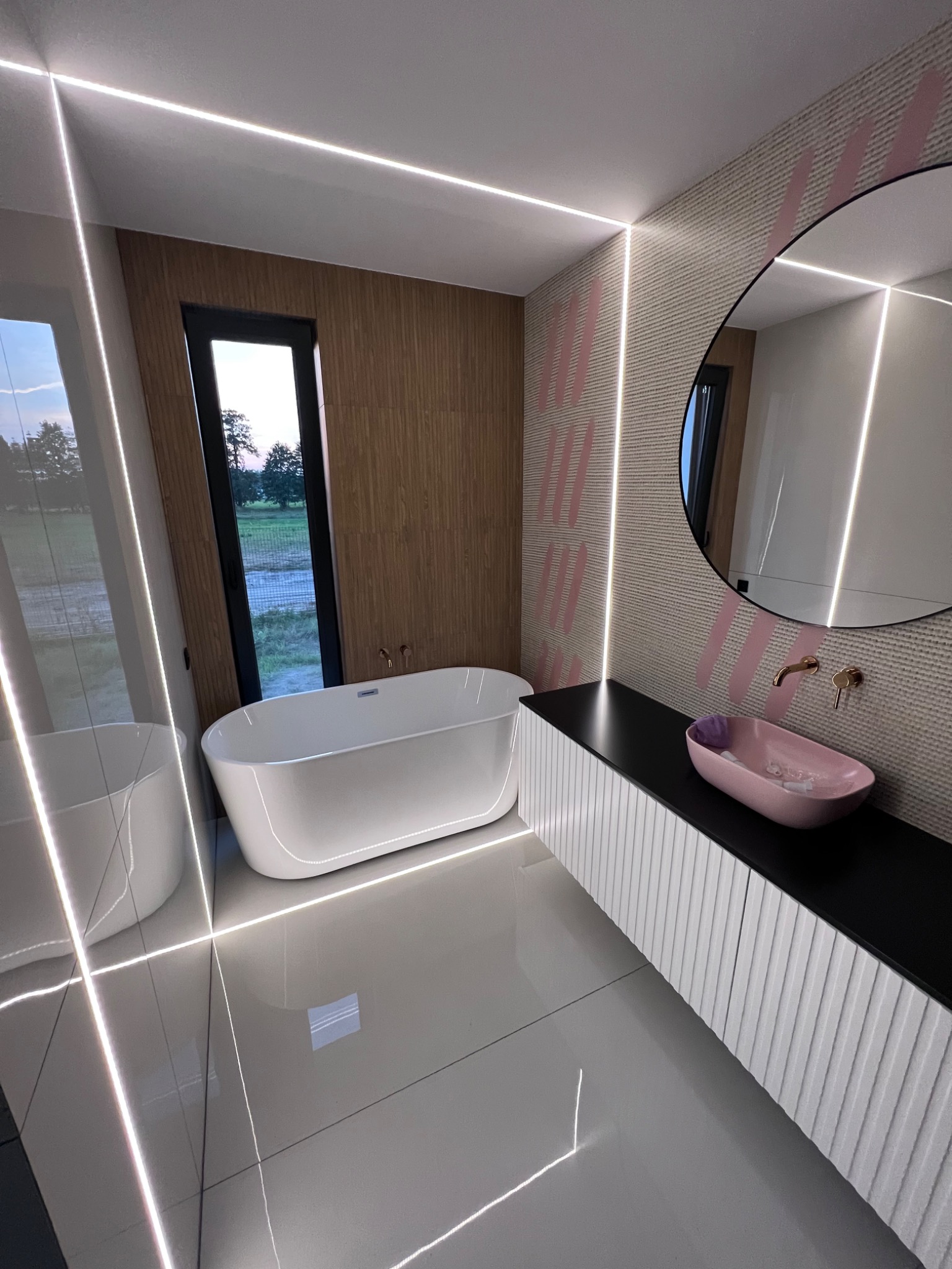 Nowoczesna łazienka z wanną wolnostojącą, okrągłym lustrem, podświetleniem LED i umywalką w odcieniach różu. Minimalistyczny design z akcentami drewna i geometrycznymi wzorami.