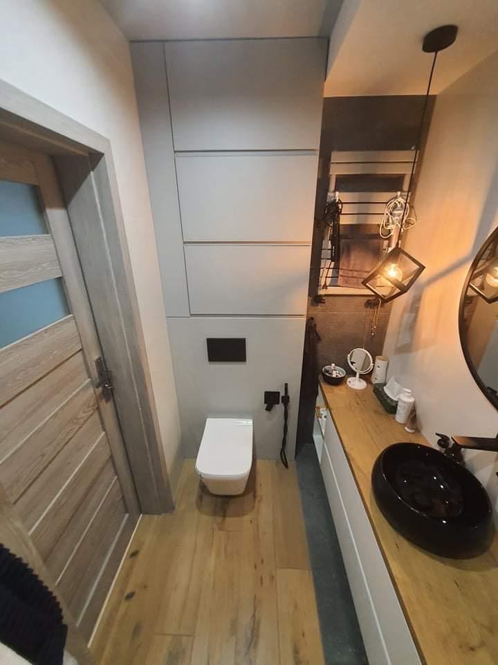 Nowoczesna aranżacja łazienki we Włocławku: drewniana podłoga, biała toaleta, szafki w zabudowie i czarna umywalka nablatowa. Minimalistyczny design i funkcjonalność.