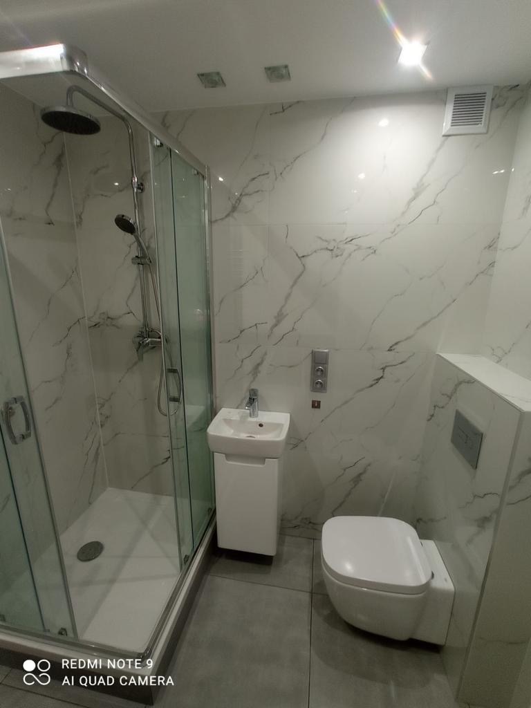 Nowoczesna łazienka z kabiną prysznicową, umywalką na szafce i podwieszaną toaletą. Białe płytki imitujące marmur na ścianach, szare płytki na podłodze.