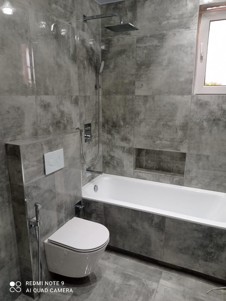 Szara łazienka z wanną, toaletą podwieszaną i deszczownicą. Płytki imitujące beton, wnęka w ścianie nad wanną, minimalistyczny styl.