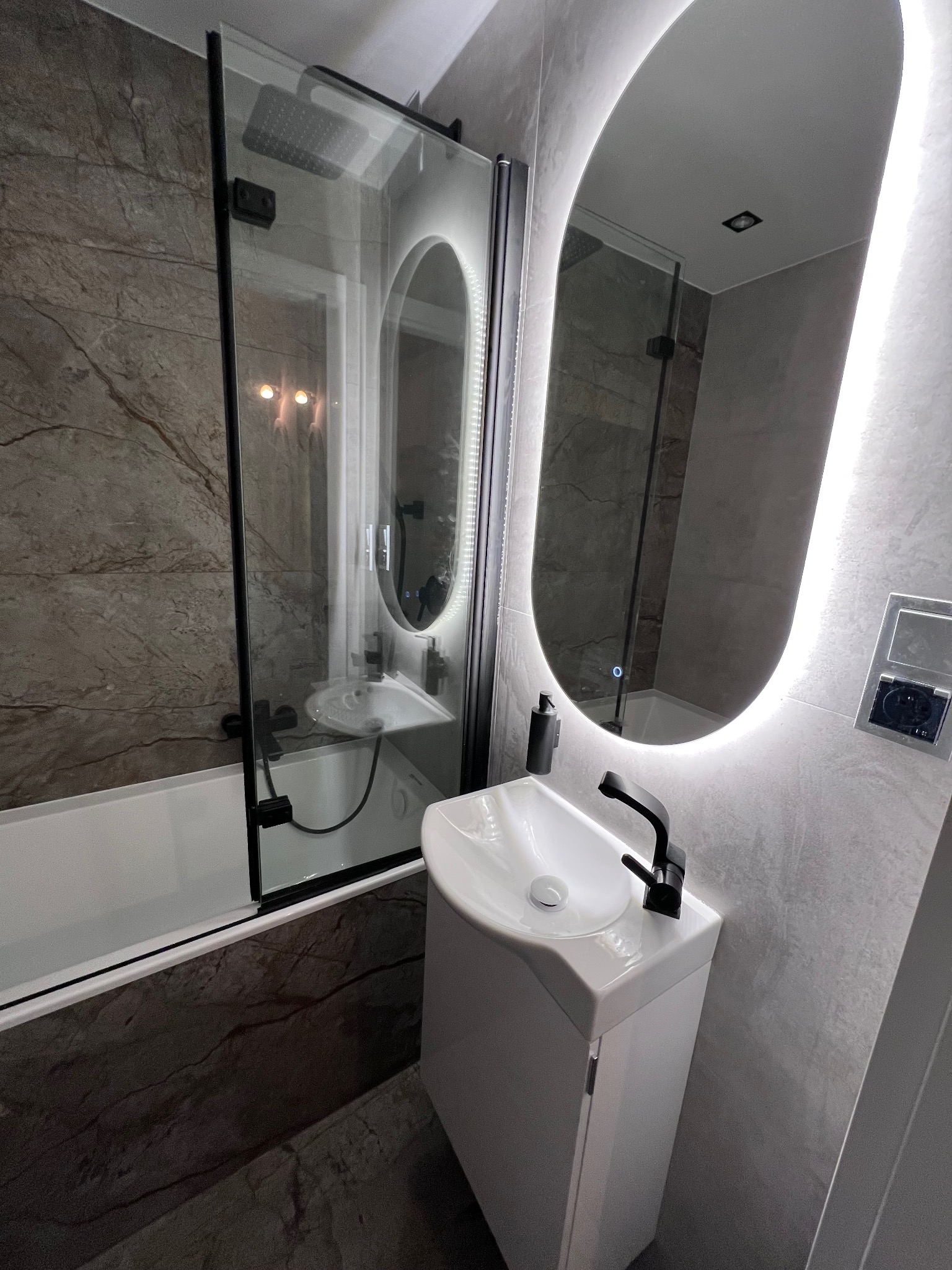 Nowoczesna łazienka z umywalką na szafce, kabiną prysznicową z czarnymi ramami i podświetlanym lustrem. Szare płytki imitujące marmur na ścianach i podłodze.