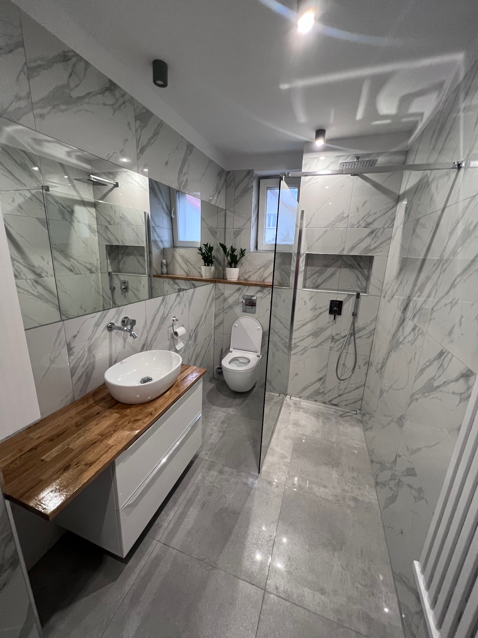 Nowoczesna łazienka z marmurowymi płytkami, drewnianym blatem pod umywalką i szklaną kabiną prysznicową typu walk-in. Minimalistyczny design i eleganckie wykończenie.