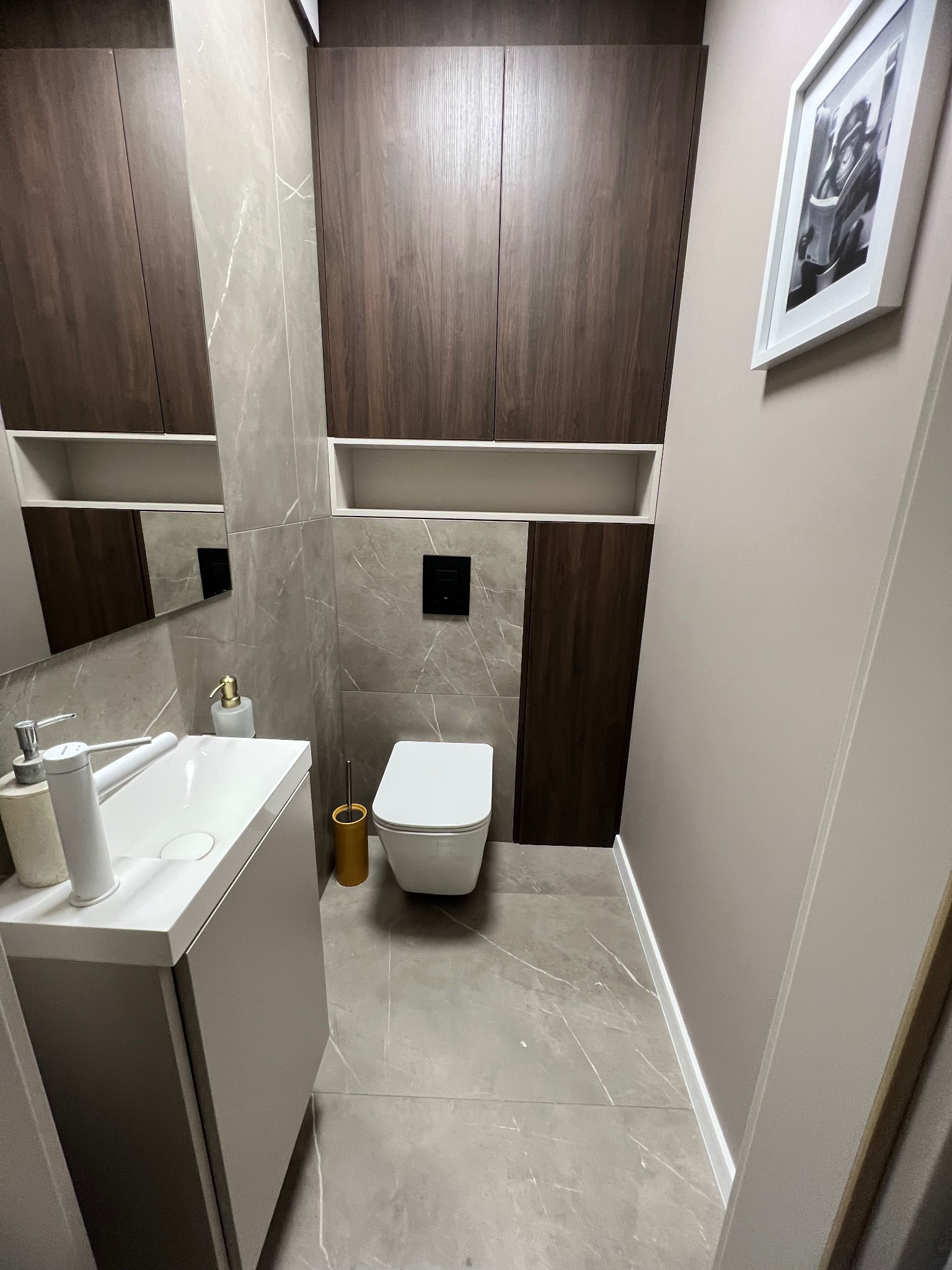 Nowoczesna łazienka w szarościach z marmurowymi płytkami, wiszącą toaletą i umywalką z szafką. Na ścianie wisi czarno-biała grafika w białej ramie.