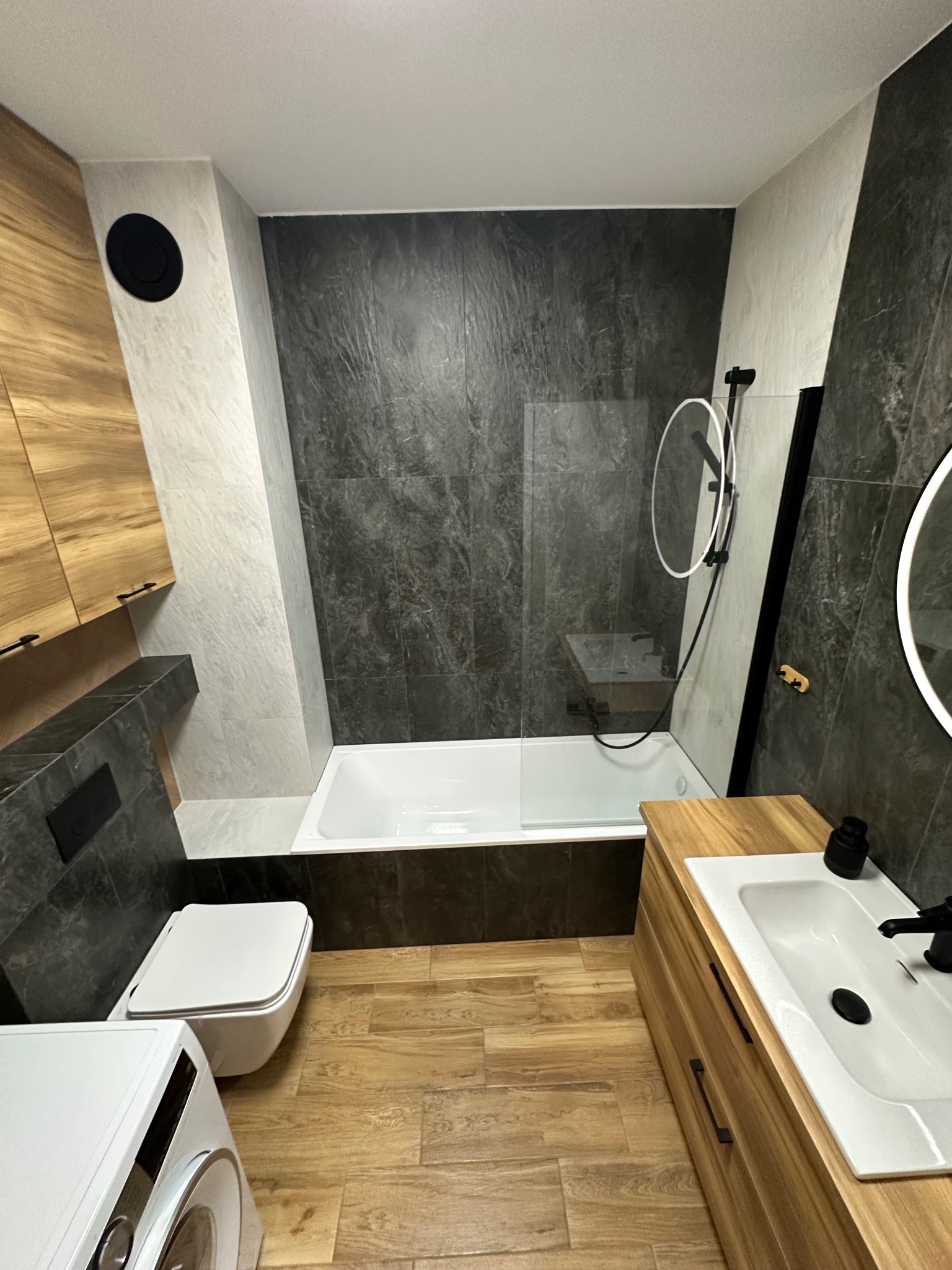 Nowoczesna łazienka z drewnianymi akcentami: szafka pod umywalką z jasnego drewna, szare płytki imitujące kamień, biała wanna z parawanem i toaleta podwieszana.