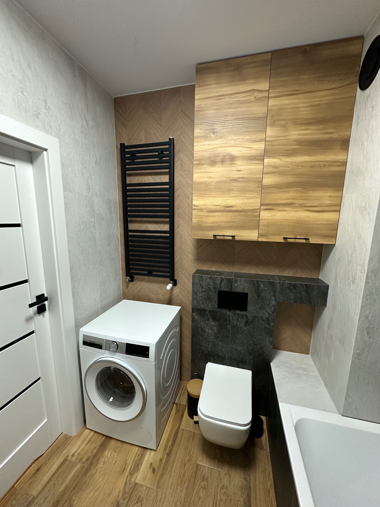 Nowoczesna łazienka po remoncie: pralka Bosch, wisząca toaleta, drewniane szafki, grzejnik drabinkowy i płytki imitujące drewno. Całość w jasnych, stonowanych barwach.