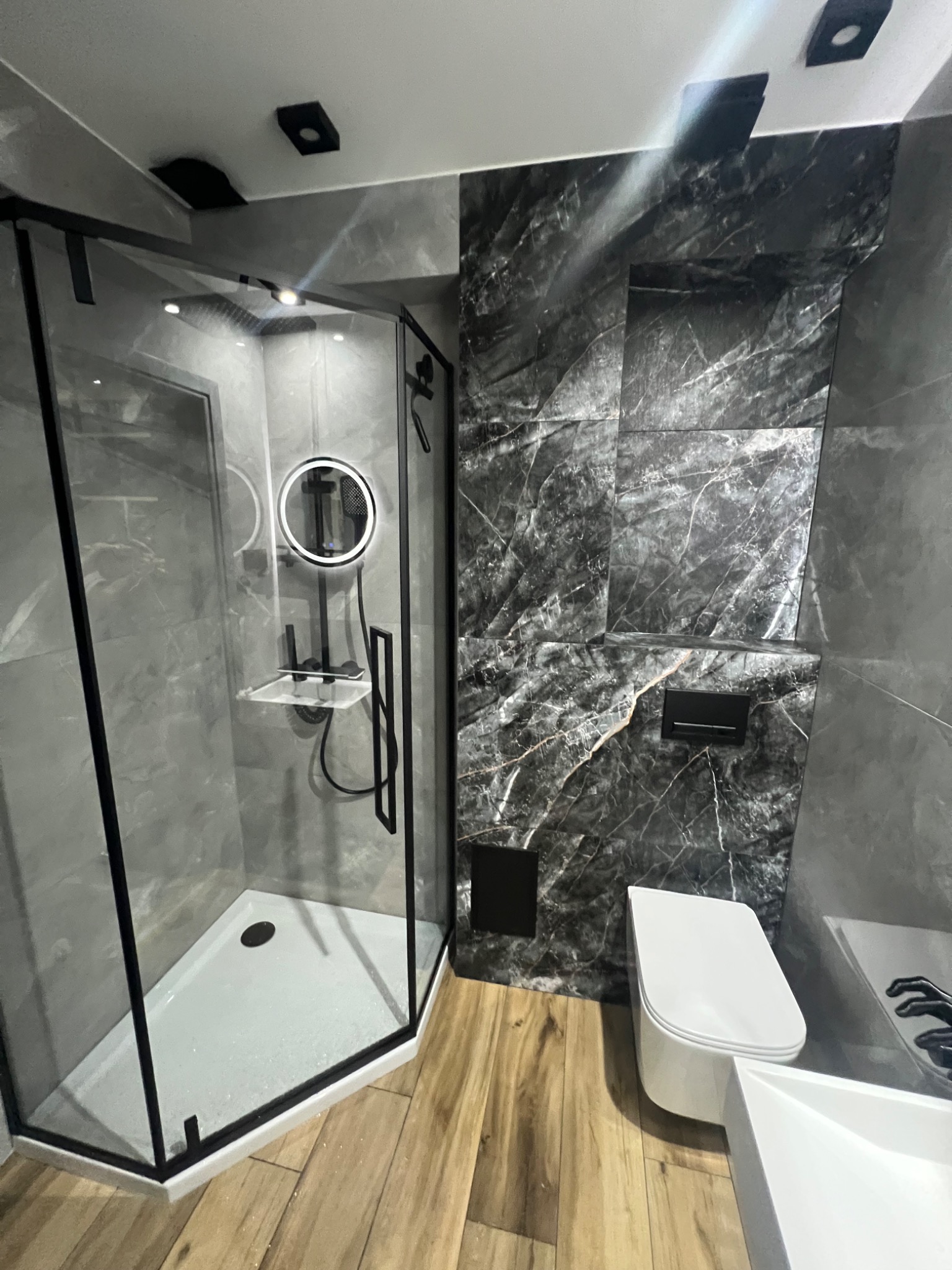 Nowoczesna łazienka z kabiną prysznicową typu walk-in, wykończona płytkami imitującymi marmur i drewnopodobną podłogą. Uwagę zwraca okrągłe lustro z podświetleniem LED.