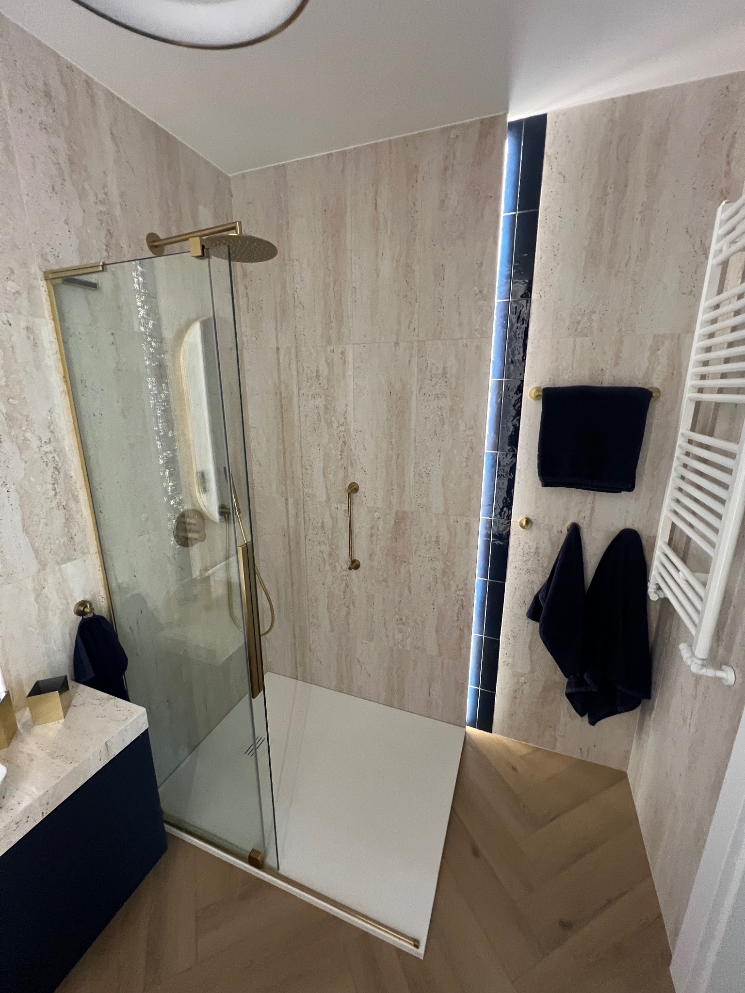 Nowoczesna łazienka z kabiną prysznicową typu walk-in, wykończona beżowymi płytkami imitującymi kamień. Złote detale armatury i czarne ręczniki dodają elegancji.