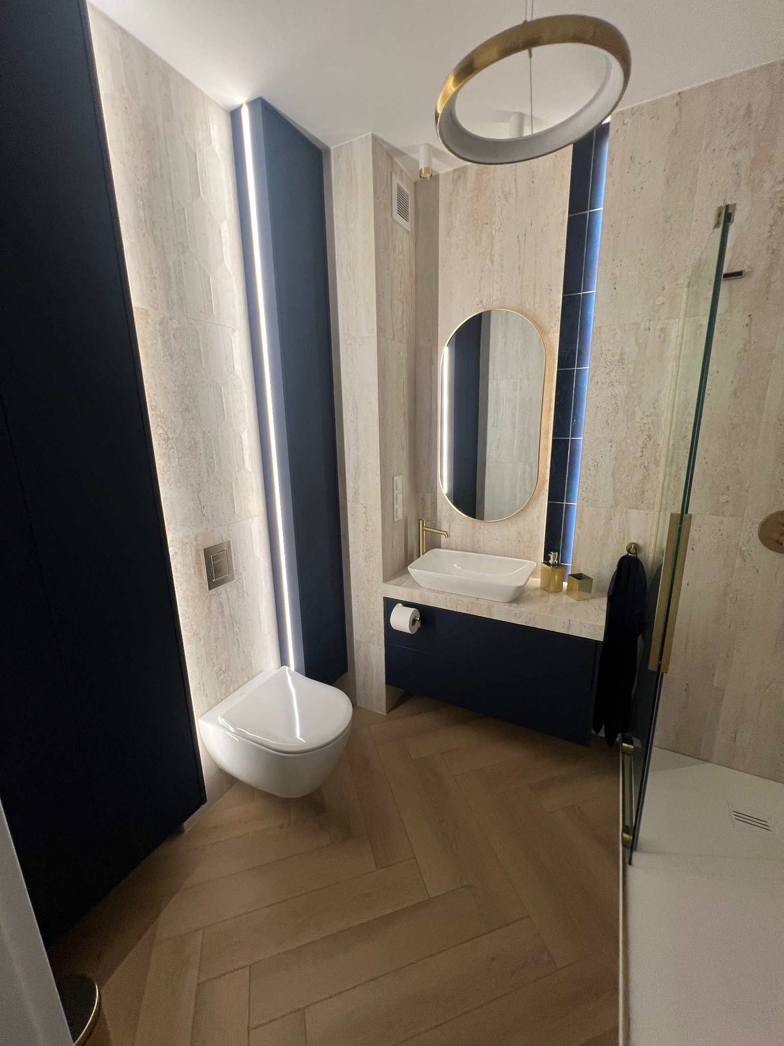 Elegancka łazienka po renowacji: beżowe płytki, drewniana podłoga w jodełkę, czarne akcenty i oświetlenie LED. Umywalka nablatowa i owalne lustro z złotą ramą.