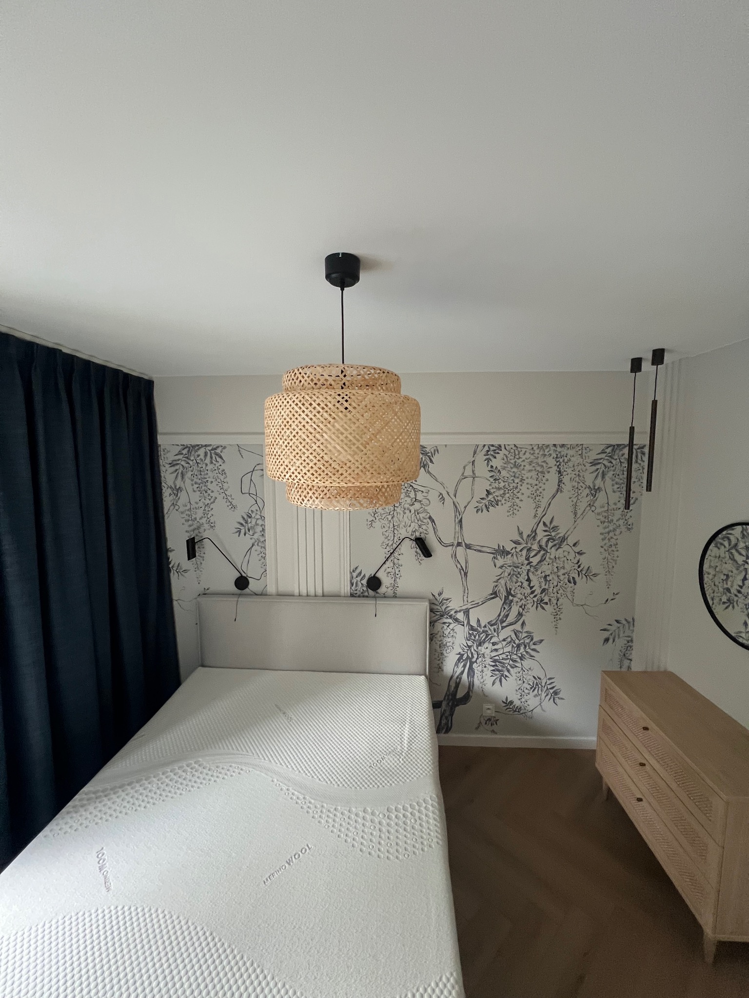 Jasna sypialnia z łóżkiem, komodą i lampą z wikliny. Na ścianie tapeta z motywem kwitnącej glicynii. Ciemne zasłony po lewej stronie kadru.