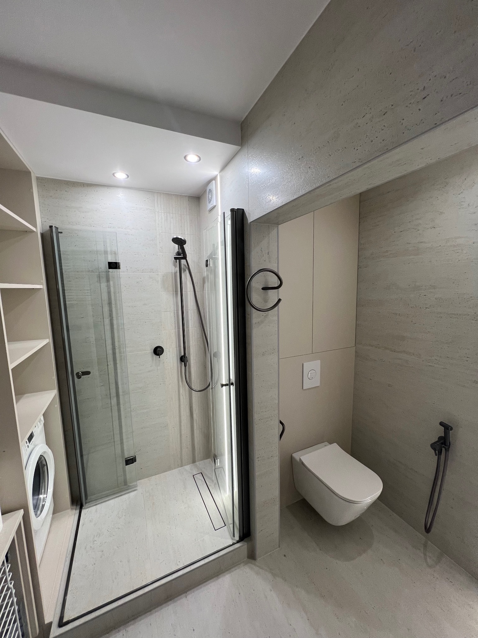 Nowoczesna łazienka z kabiną prysznicową typu walk-in, toaletą podwieszaną i pralką w zabudowie. Użyto płytek imitujących kamień w jasnych odcieniach szarości.