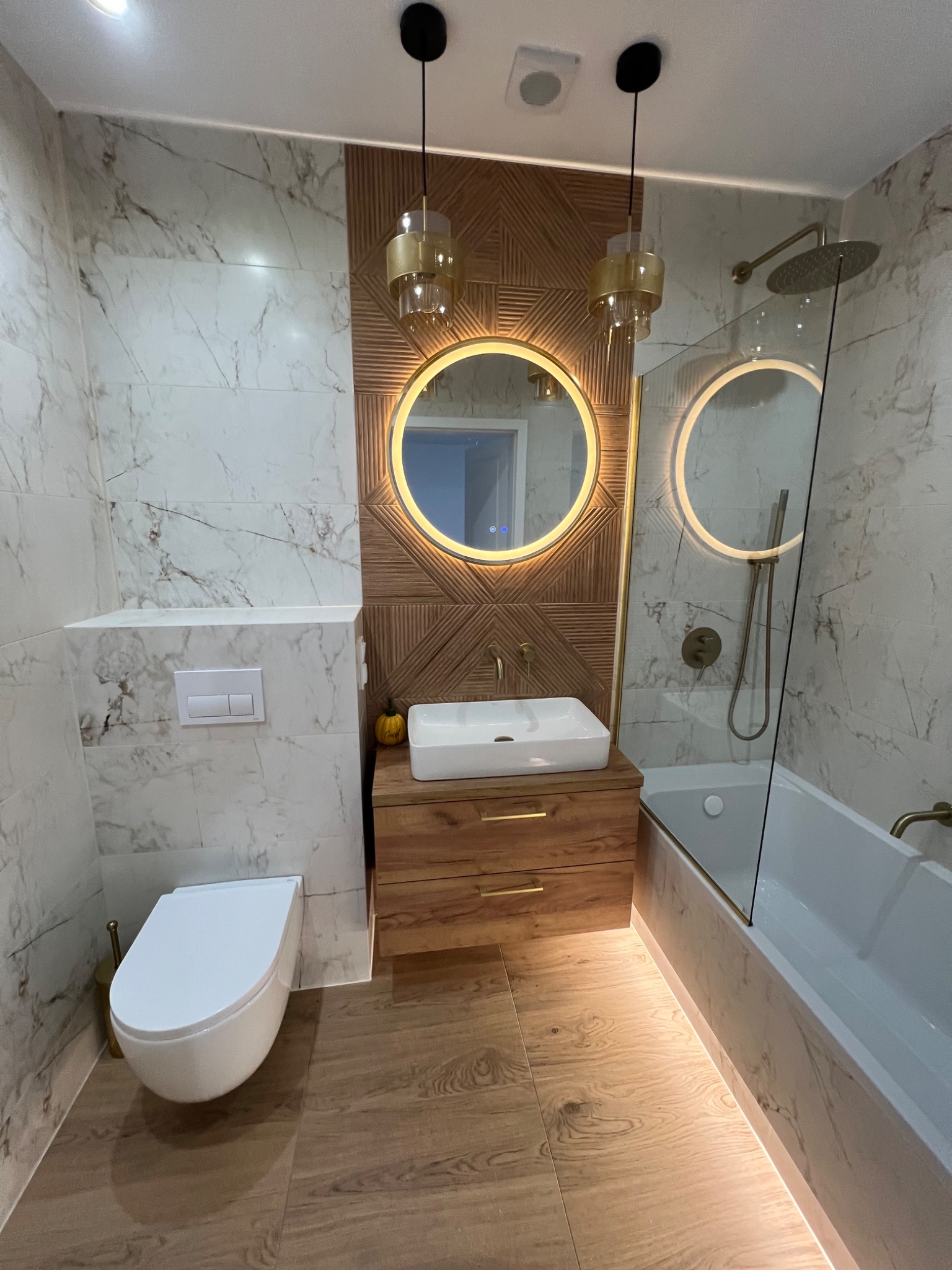 Nowoczesna łazienka z marmurowymi płytkami, drewnianymi akcentami i okrągłymi lustrami z podświetleniem LED. Uwagę przykuwają złote lampy wiszące i drewniana mozaika na ścianie.