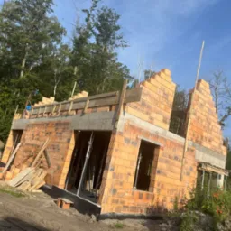 Wznoszenie ścian domu z czerwonej cegły. Widoczne szalunki i podpory. W tle drzewa i błękitne niebo. Częściowo zarysowany stos palet i desek.