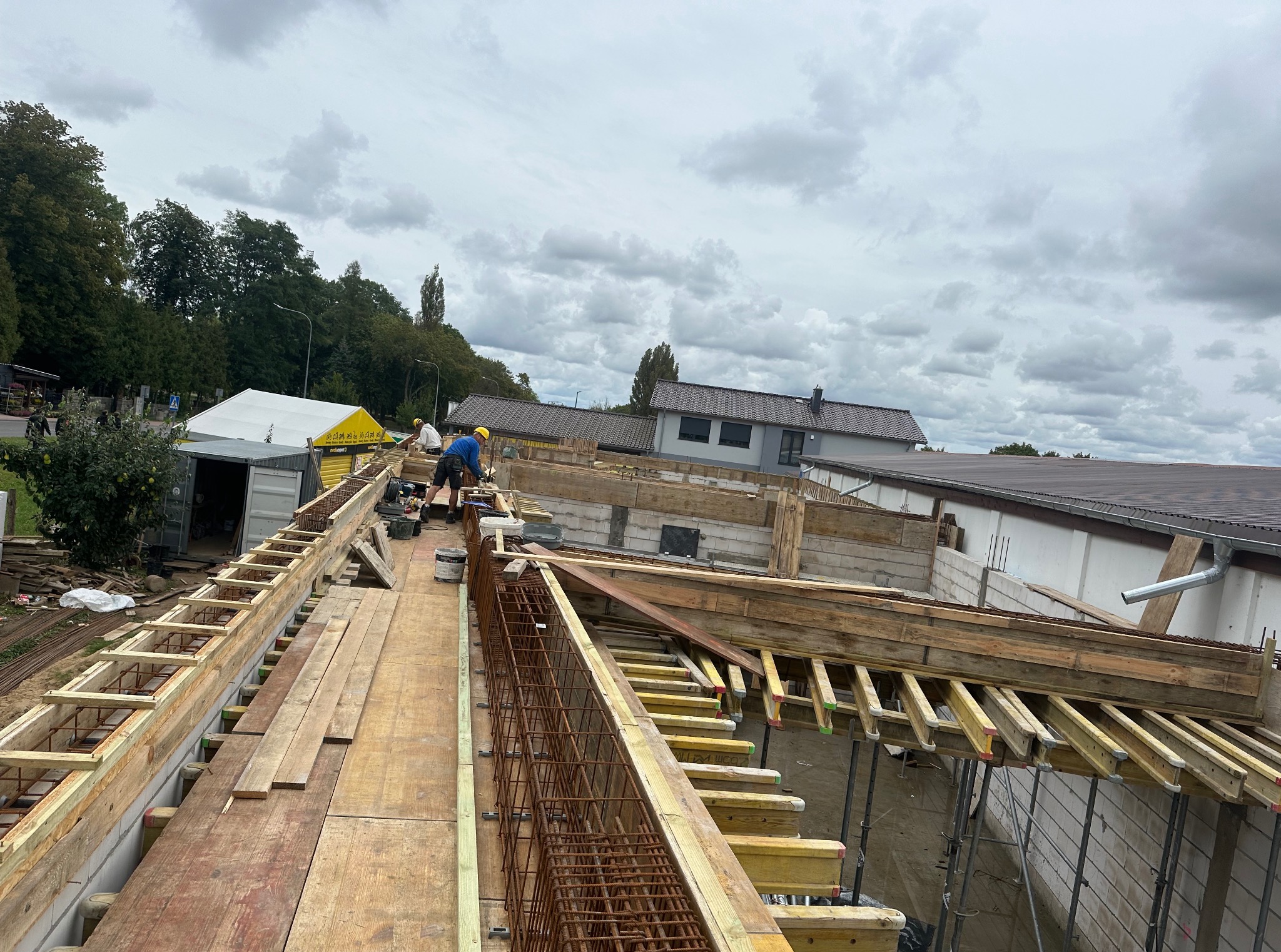 Budowa domu: zbrojenie i szalunki ścian fundamentowych z widocznymi pracownikami w kaskach, przygotowane do zalewania betonem na tle pochmurnego nieba.