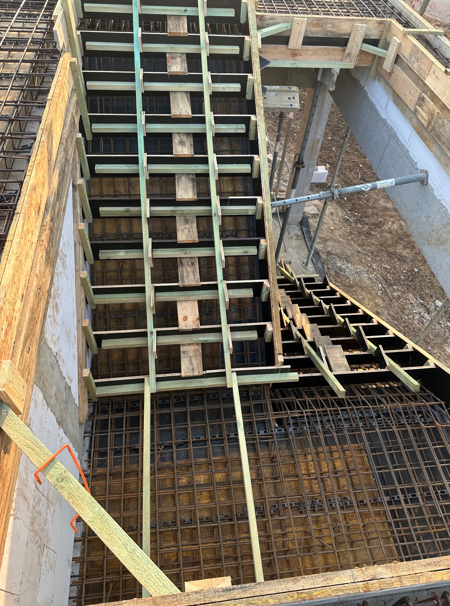 Zbrojenie schodów z desek i metalowych prętów, widok z góry na plac budowy. Elementy konstrukcyjne podtrzymywane przez podpory.
