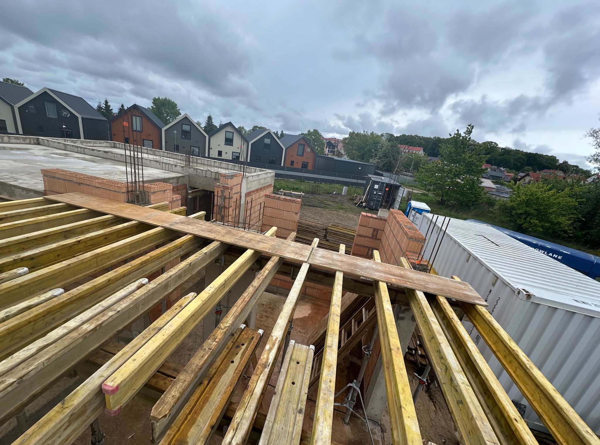 Budowa domu: widok z góry na konstrukcję drewnianą stropu, z widocznymi elementami ścian z cegły i w tle rząd nowoczesnych domów jednorodzinnych pod pochmurnym niebem.