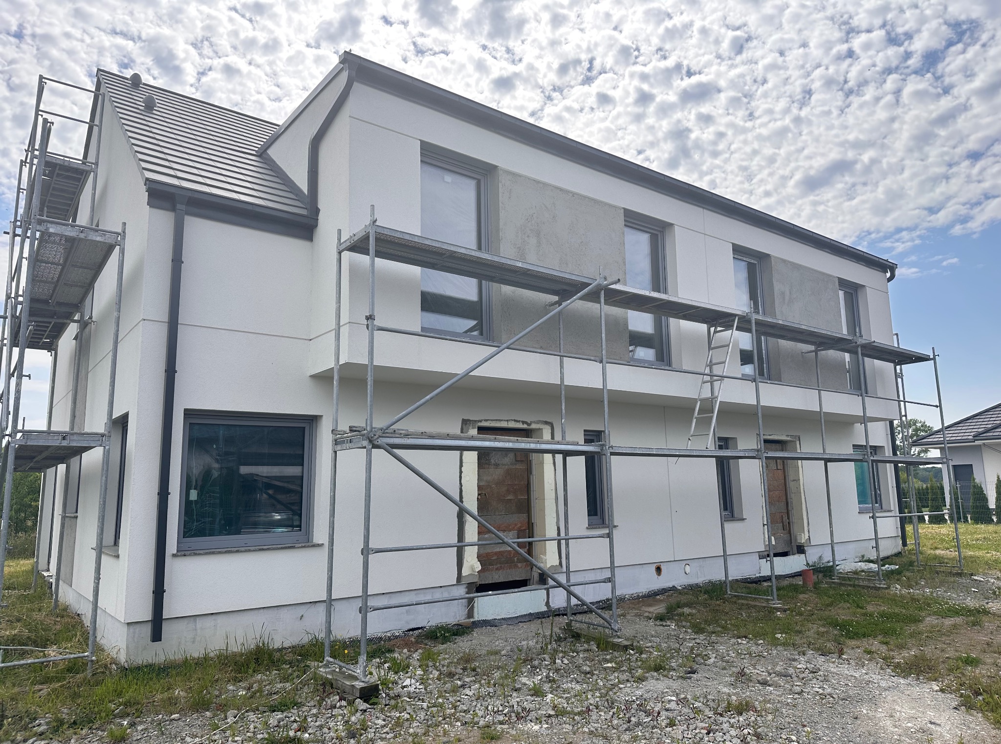Nowoczesny dom w trakcie ocieplania elewacji, widoczne rusztowania i fragmenty otynkowanej ściany. Jasna fasada kontrastuje z szarą dachówką i błękitnym niebem.