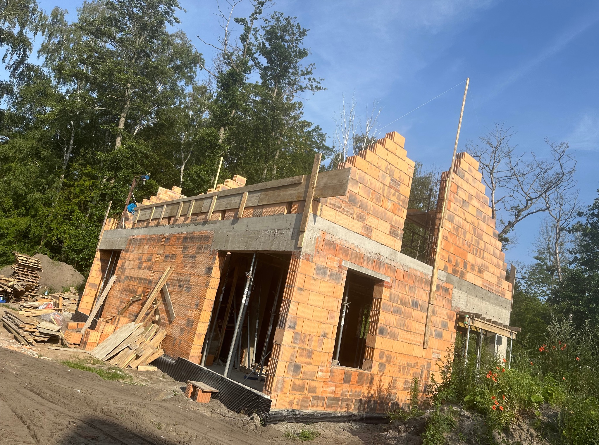 Wznoszenie ścian domu z czerwonej cegły. Widoczne szalunki i podpory. W tle drzewa i błękitne niebo. Częściowo zarysowany stos palet i desek.