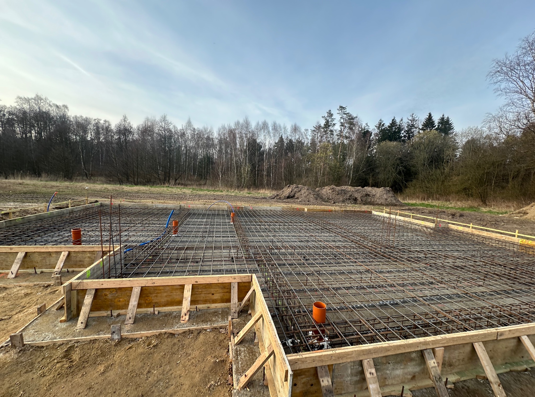 Zbrojenie fundamentów pod budowę domu jednorodzinnego na działce z widokiem na las. Drewniane szalunki i stalowe pręty tworzą siatkę. Widoczne pomarańczowe rury.