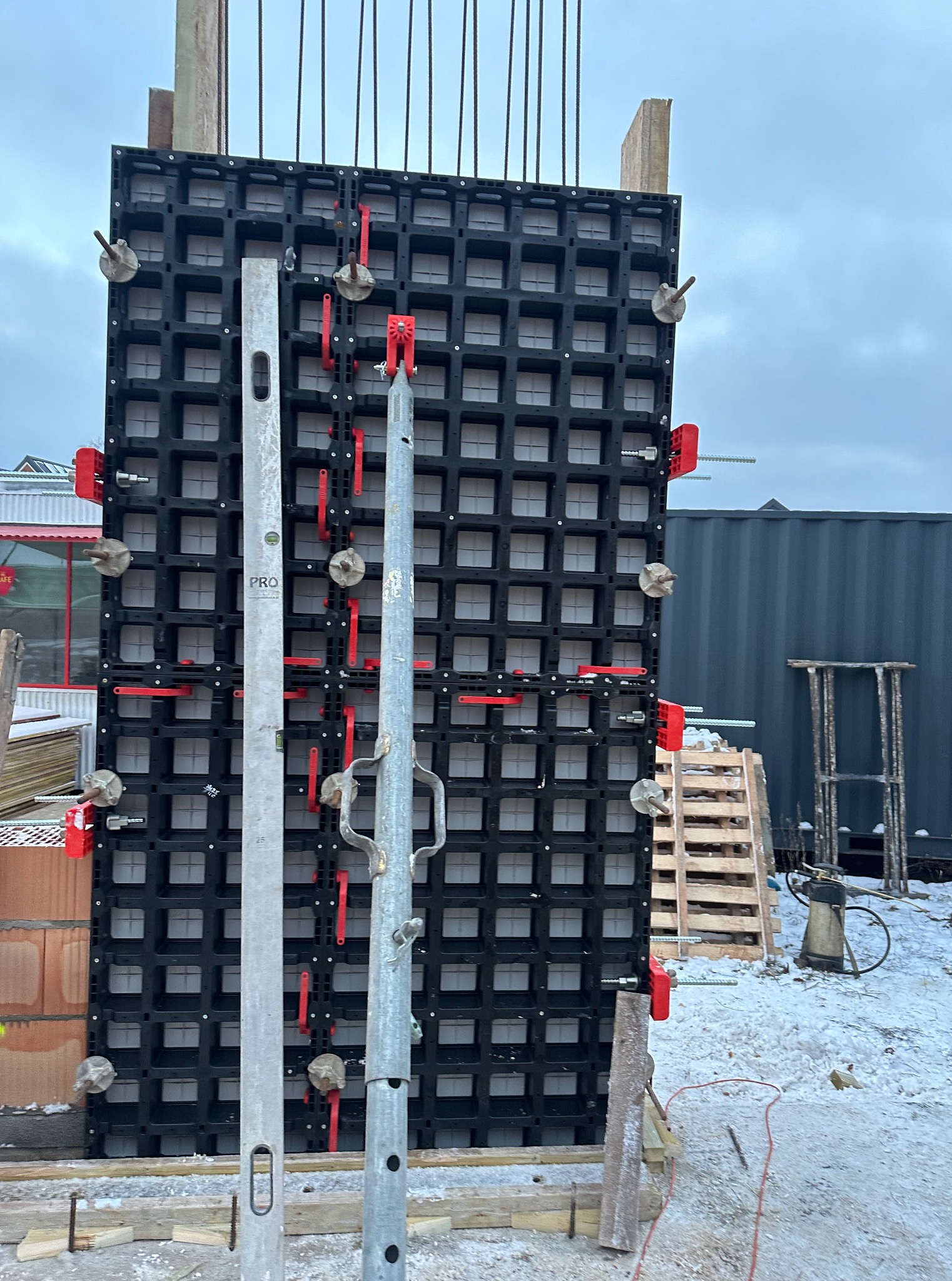 Częściowe ujęcie szalunku z tworzywa, z czerwonymi elementami mocującymi i poziomnicą, przygotowanego do zalewania betonem. Widoczne zbrojenia i zimowy krajobraz w tle.