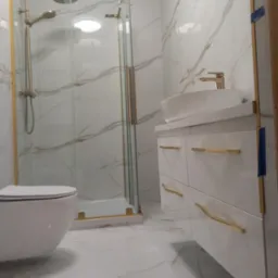 Nowoczesna łazienka z kabiną prysznicową typu walk-in z złotymi akcentami, białą umywalką nablatową i marmurowymi płytkami na ścianach i podłodze.