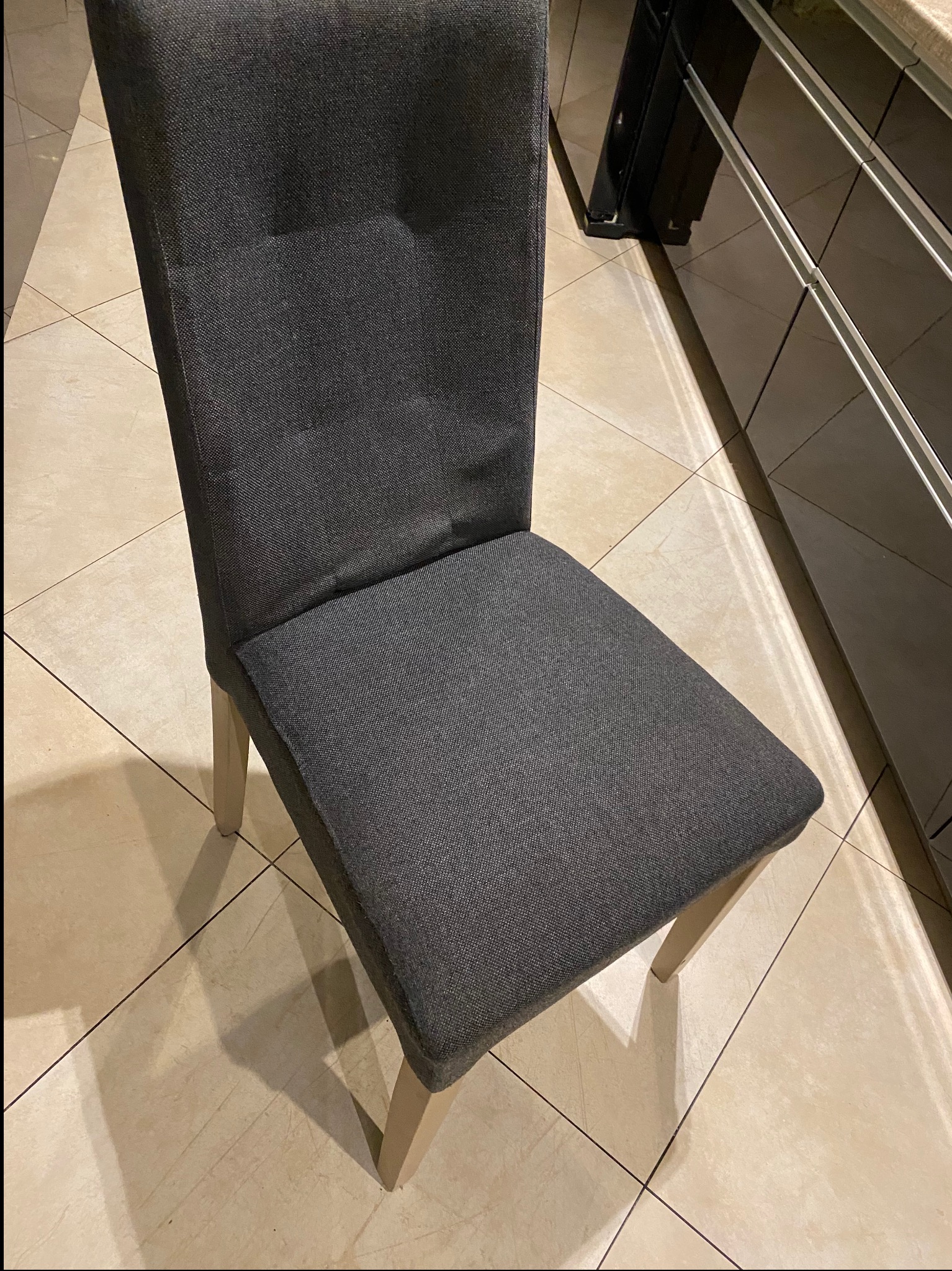 Szare krzesło tapicerowane z jasnymi nogami na tle płytek podłogowych i czarnych szafek kuchennych. Widok z góry, oświetlenie naturalne.