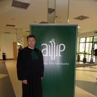 Adwokat w todze na tle zielonego baneru z logo kancelarii w jasnym, nowoczesnym wnętrzu. Widoczne elementy architektoniczne i oświetlenie.