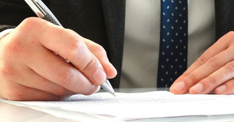 Ręka w garniturze pisze srebrnym długopisem na białych dokumentach, widoczny fragment krawata w granatowe kropki. Jasne oświetlenie, neutralne tło.