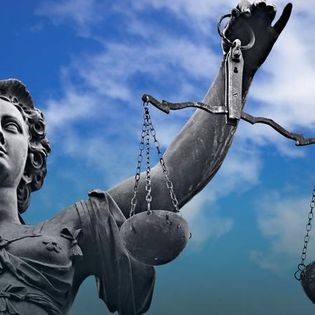 Posąg Temidy, bogini sprawiedliwości, na tle błękitnego nieba. Statua z wagą i mieczem, symbolizuje bezstronność i prawo. Ujęcie od dołu podkreśla powagę.