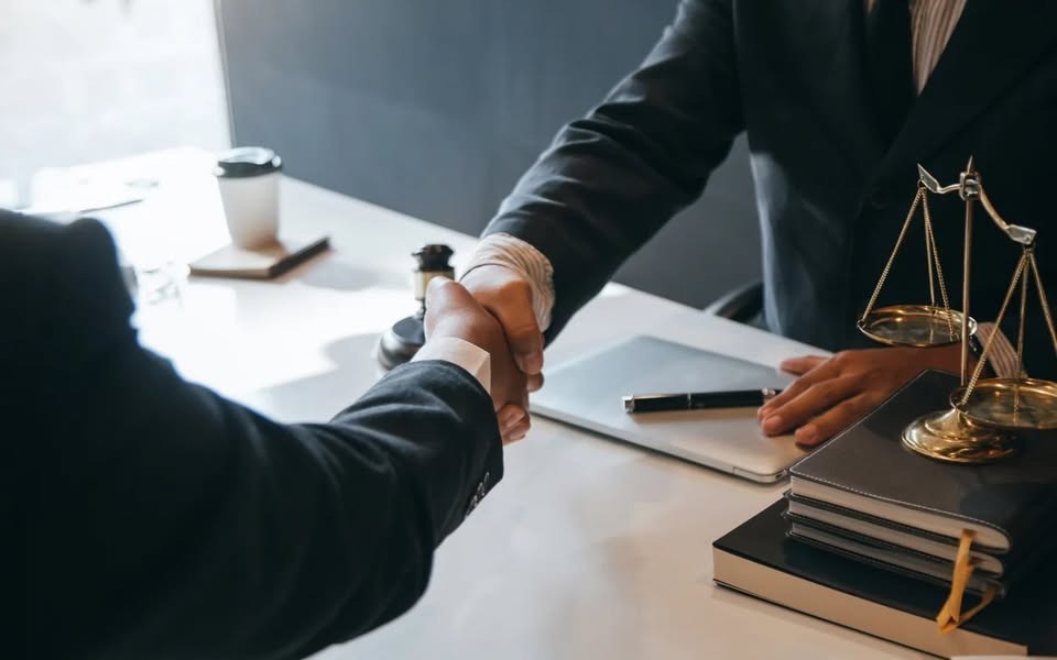 Uścisk dłoni nad biurkiem z wagą Temidy, laptopem i notatnikami. Symbol zgody i profesjonalizmu w sprawach prawnych.