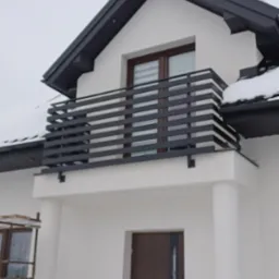 Nowoczesny balkon z poziomą, czarną balustradą na tle białej elewacji domu w zimowej scenerii. Widoczne detale wykończenia i elementy konstrukcyjne.