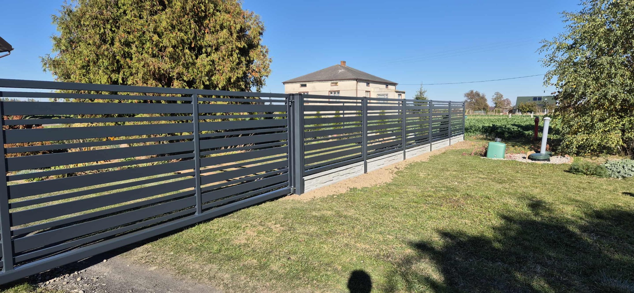 Nowoczesne ogrodzenie metalowe w kolorze grafitowym z poziomymi, szerokimi szczeblami, na tle zielonego trawnika i domu. Słoneczny dzień.