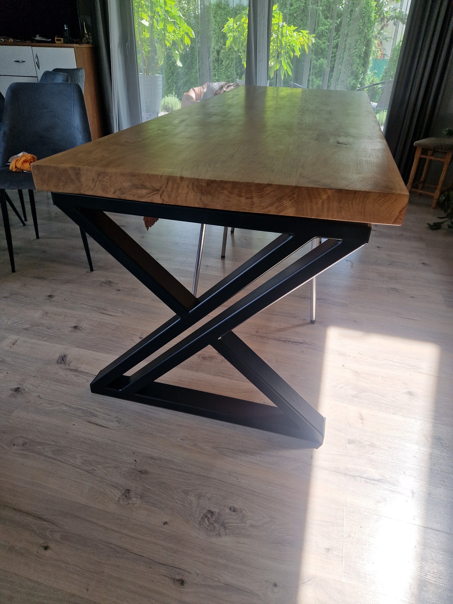 Stół z blatem z jasnego drewna i czarną, metalową podstawą o geometrycznym kształcie na tle okna z widokiem na zieleń. Nowoczesny design.