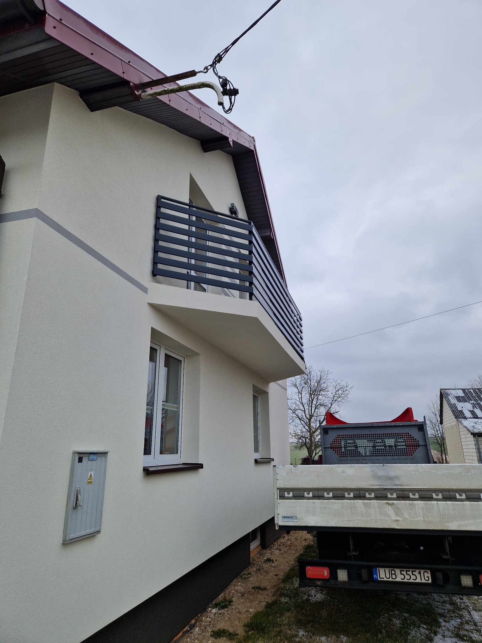 Elegancka, pozioma balustrada balkonowa z czarnych metalowych profili, zamontowana na balkonie domu o jasnej elewacji, widziana z boku. Na tle pochmurne niebo.