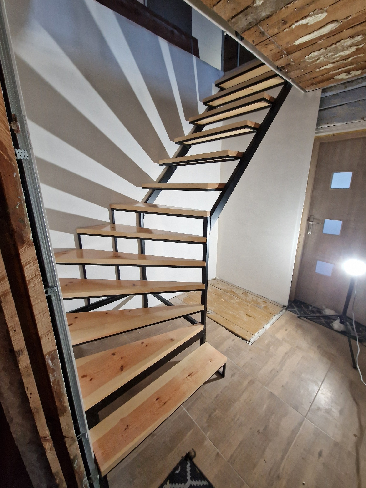 Nowoczesne schody metalowe z drewnianymi stopniami w trakcie montażu, widoczne elementy konstrukcyjne i surowe wykończenie wnętrza, oświetlenie robocze.