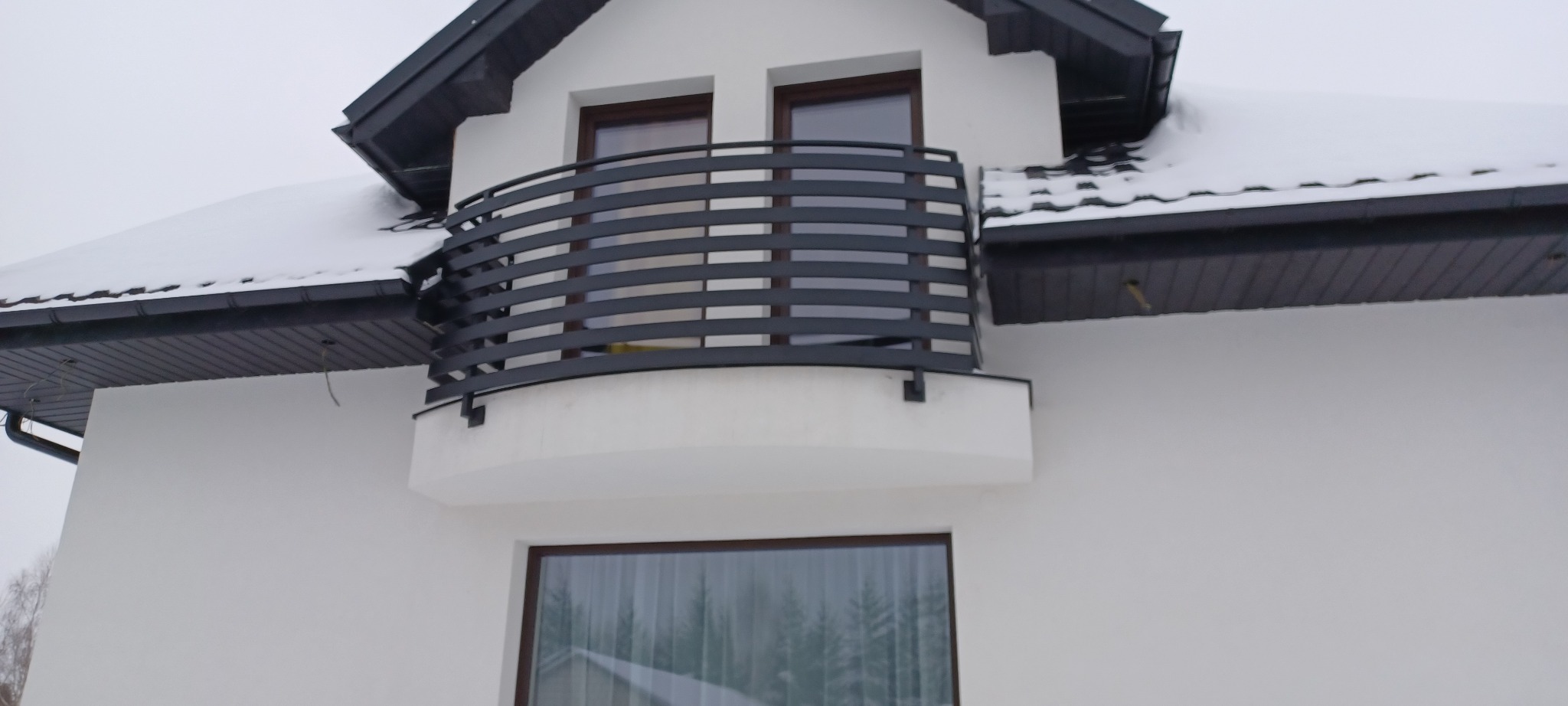 Nowoczesna, czarna balustrada balkonowa z poziomych profili na białym budynku w zimowej scenerii. Minimalistyczny design i śnieg na dachu.