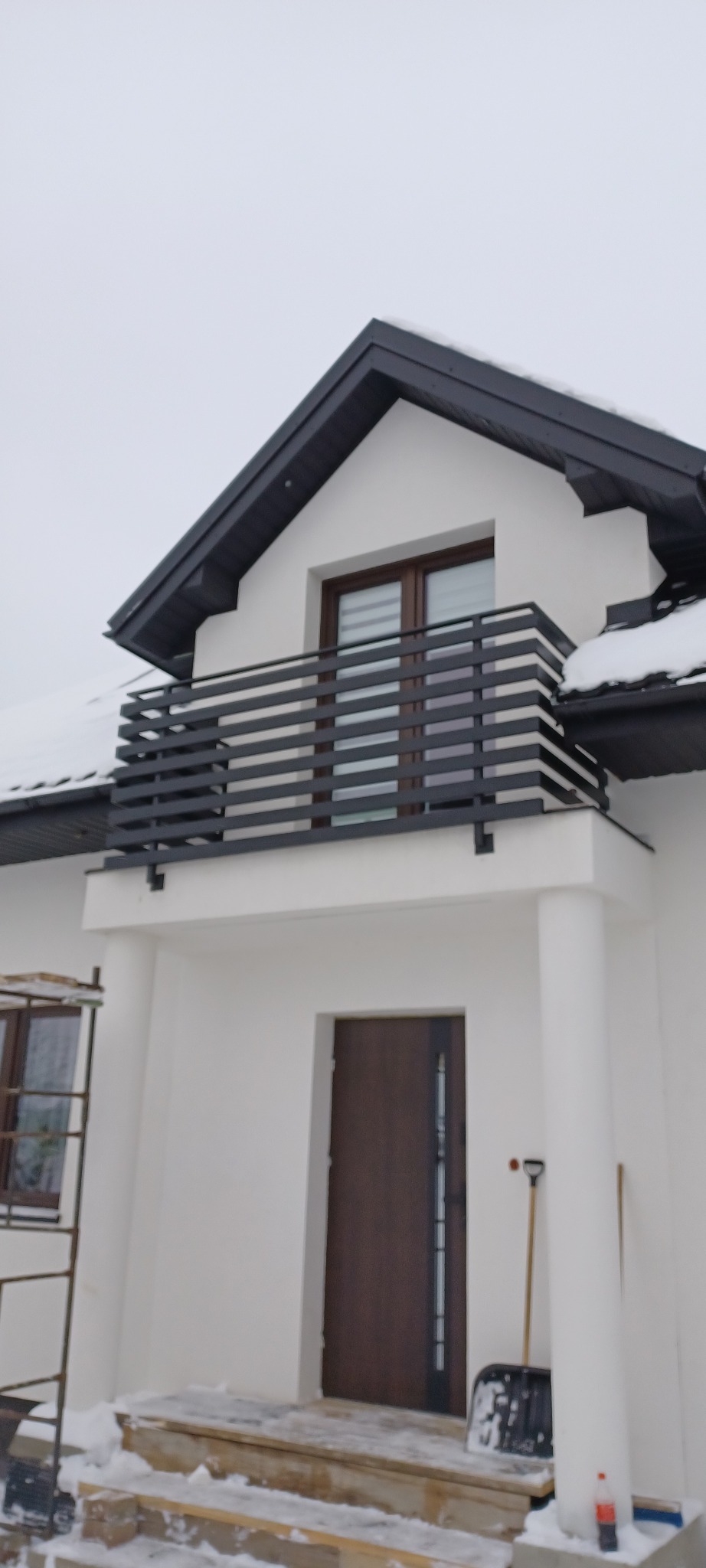 Nowoczesny balkon z poziomą, czarną balustradą na tle białej elewacji domu w zimowej scenerii. Widoczne detale wykończenia i elementy konstrukcyjne.