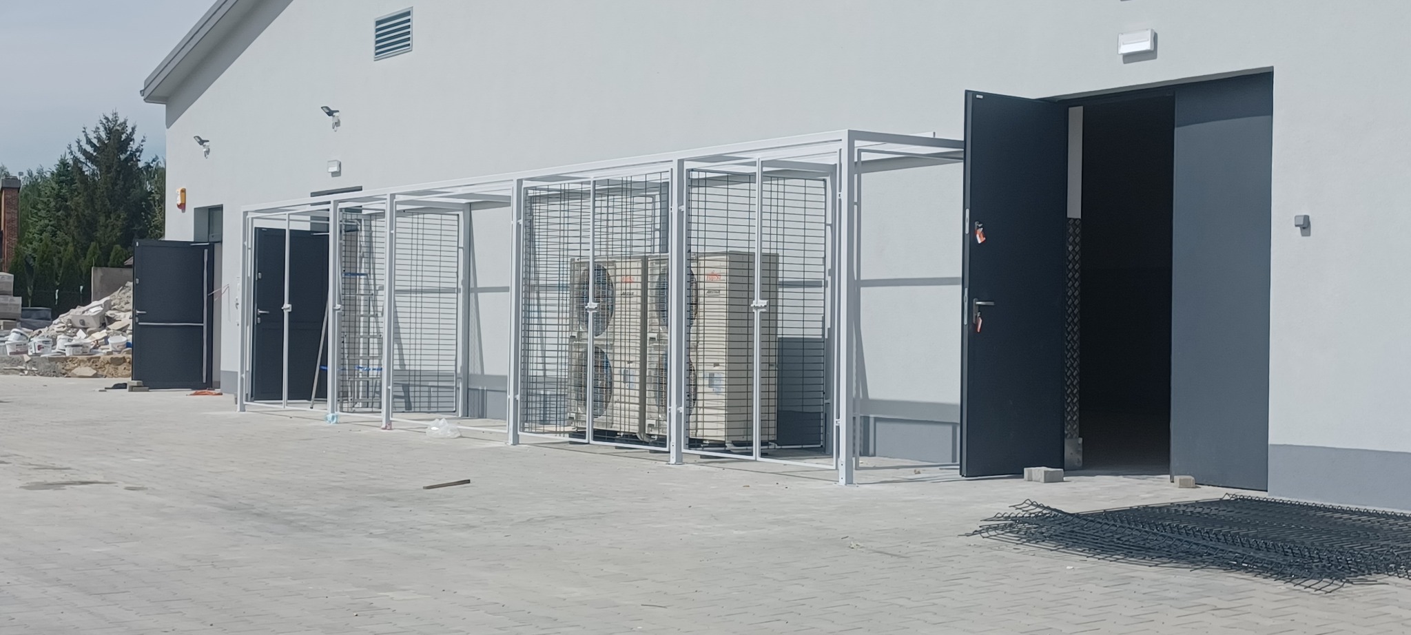Metalowa klatka na klimatyzatory, wykonana w Bychawie. Konstrukcja z siatki i profili, pomalowana na biało, chroni urządzenia przed uszkodzeniami i kradzieżą. Widok z zewnątrz.