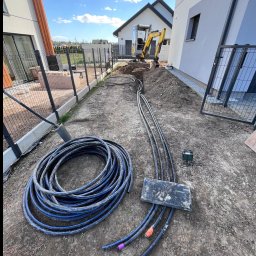 KAROL KOT - Instalacja rur z kolorowymi końcówkami w ziemi, obok zwój rur i minikoparka na tle zabudowy jednorodzinnej. Prace ziemne w toku.