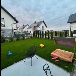 KAROL KOT - Zadbany trawnik przed nowoczesnymi domami w zabudowie szeregowej. Widoczne zabawki dziecięce, krzesło i piaskownica. Regularne nasadzenia drzew i krzewów.