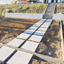 Układanie ścieżki z płyt na gruncie, widoczne obrzeża i schody w tle. Prace ziemne przygotowujące teren pod przyszły ogród.