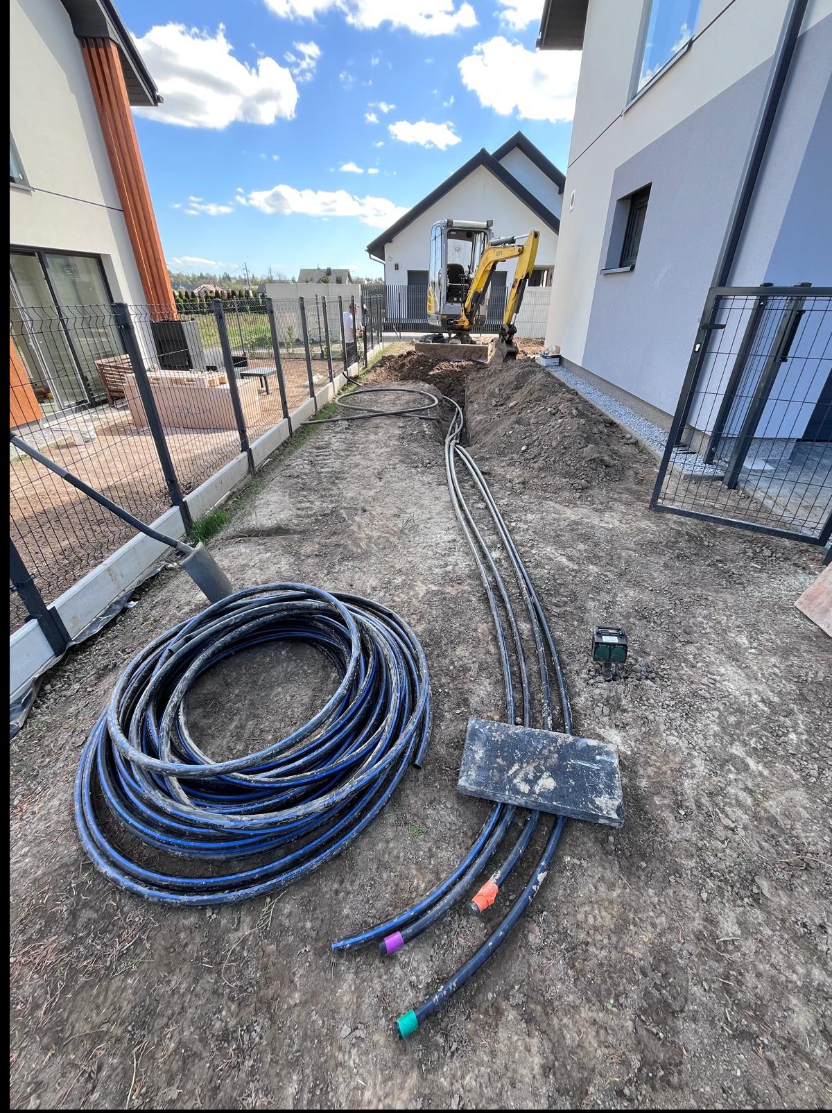 Instalacja rur z kolorowymi końcówkami w ziemi, obok zwój rur i minikoparka na tle zabudowy jednorodzinnej. Prace ziemne w toku.