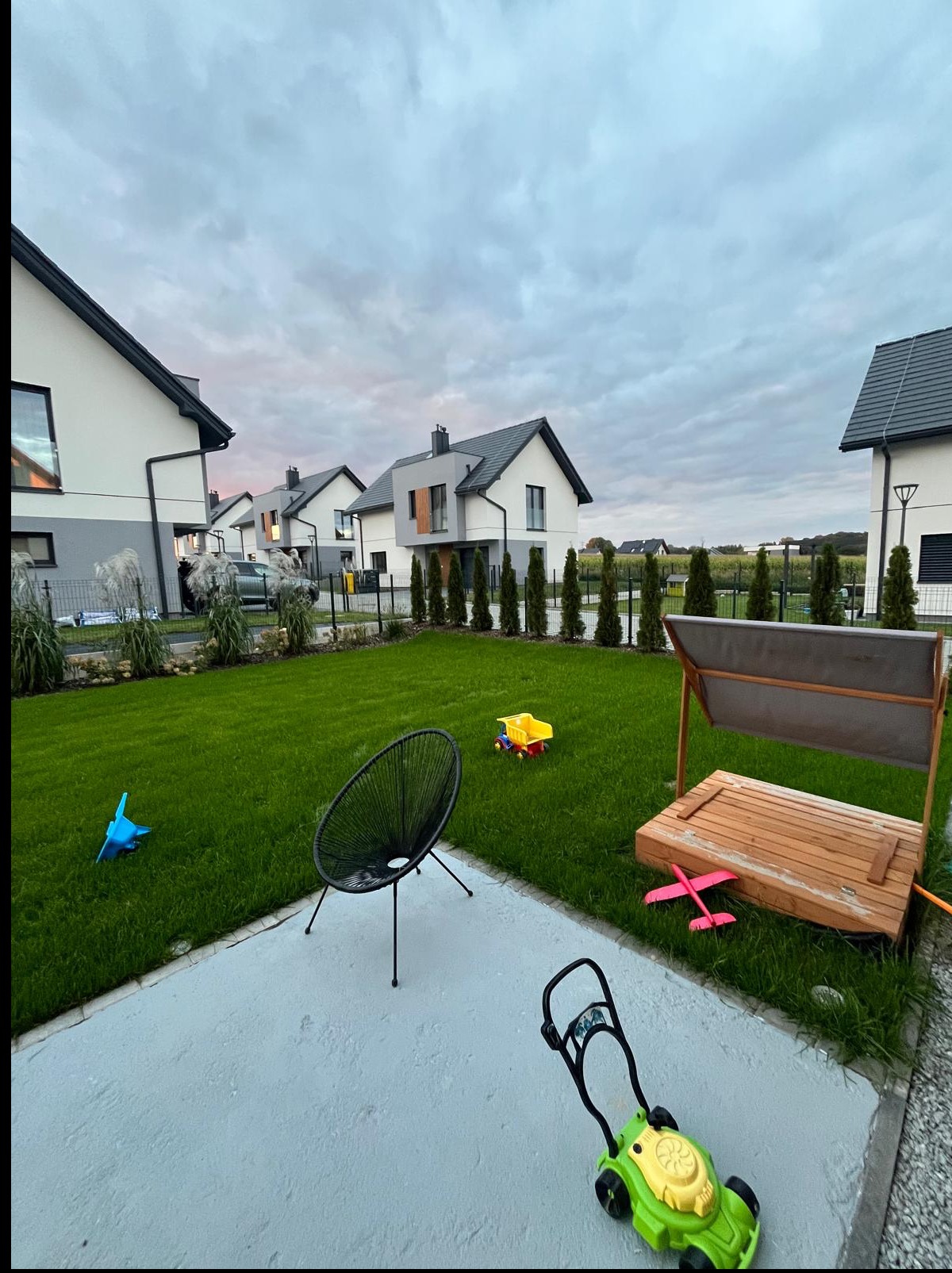Zadbany trawnik przed nowoczesnymi domami w zabudowie szeregowej. Widoczne zabawki dziecięce, krzesło i piaskownica. Regularne nasadzenia drzew i krzewów.