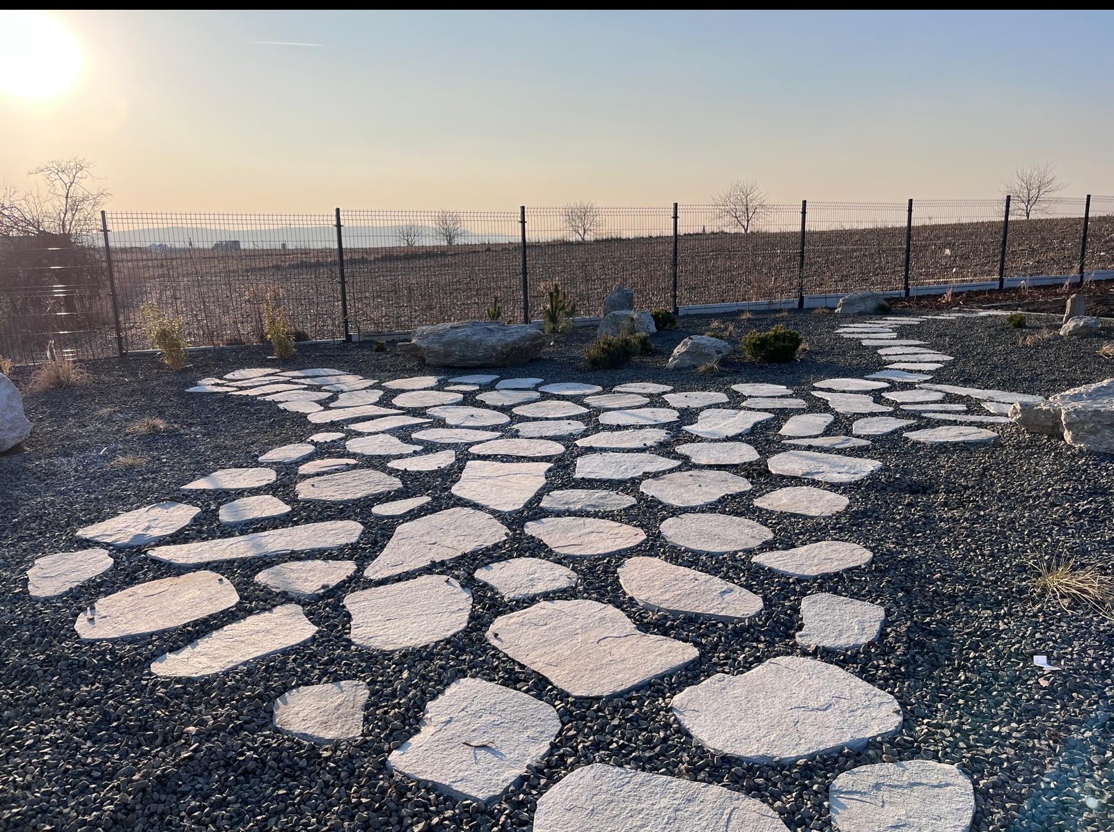 Ścieżka z nieregularnych płyt kamiennych na żwirowym podłożu, prowadząca przez ogród z młodymi drzewkami i ozdobnymi kamieniami, ogrodzony siatką.