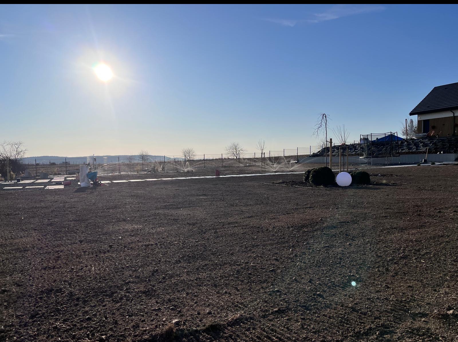 Nowo założony ogród z uruchomionym systemem nawadniania, widoczne zraszacze. W tle dom i błękitne niebo z promieniami słońca. Świeżo przygotowana ziemia pod zasiew trawy.