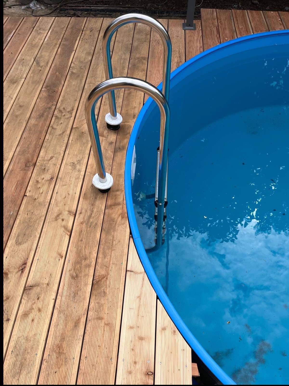 Niebieski basen ogrodowy z metalową drabinką, otoczony drewnianym tarasem. Woda w basenie odbija otaczające drzewa i niebo, tworząc spokojną atmosferę.