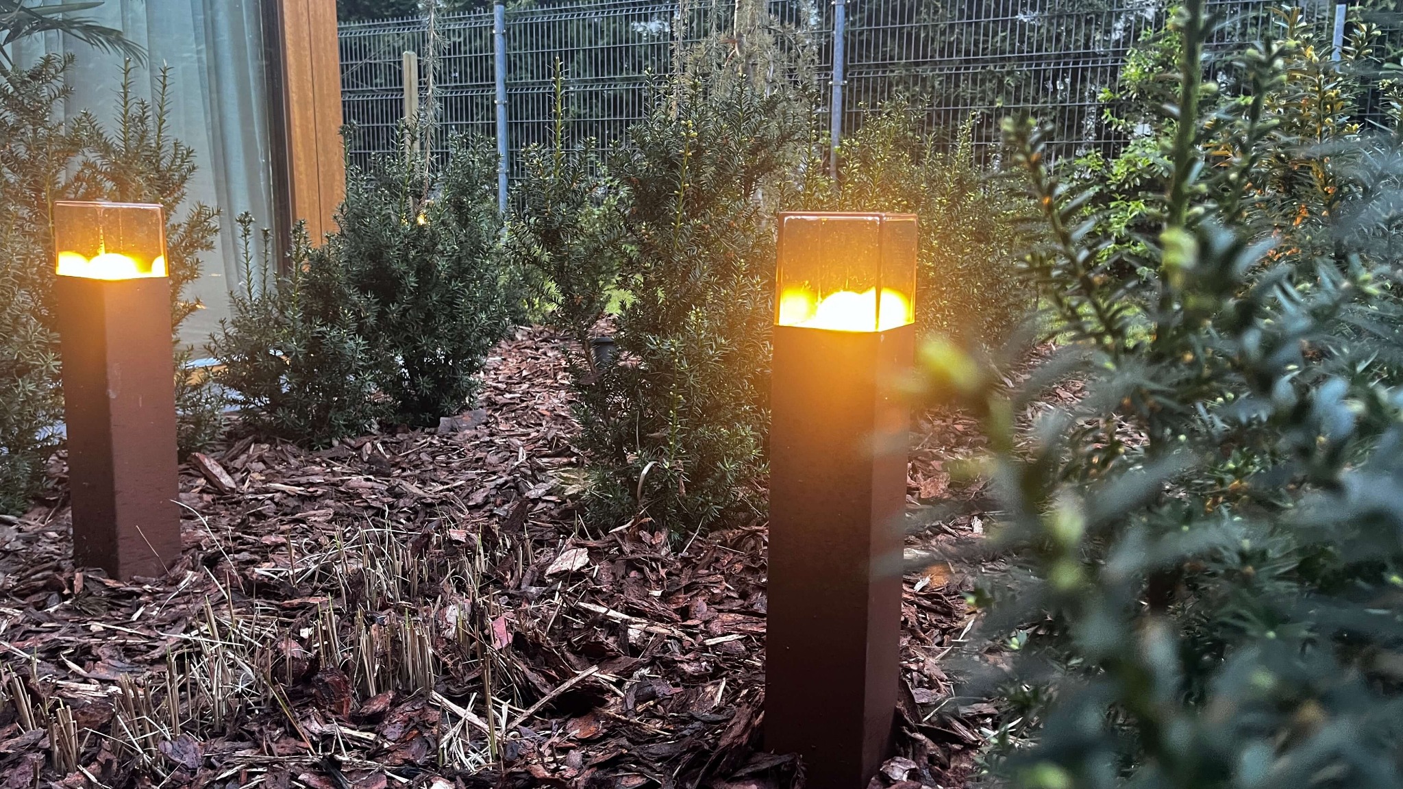 Oświetlenie ogrodu: Dwie nowoczesne, kwadratowe lampy ogrodowe z ciepłym światłem, otoczone korą i iglakami. Metalowy płot w tle, wieczorny klimat.